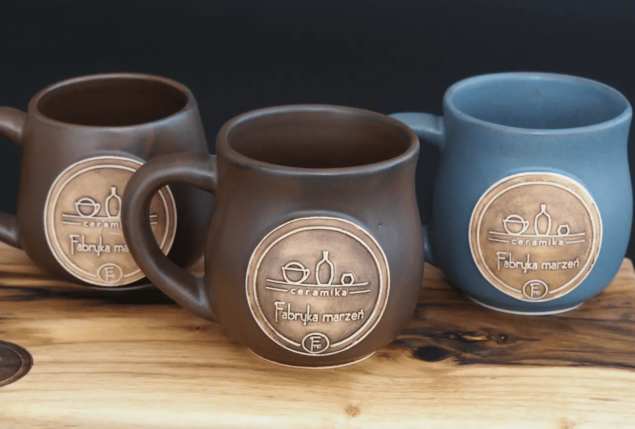 3 large and neat looking mugs of the fmceramics brand engraved with the logo of this brand, the mugs are arranged next to each other on a wooden table, they are in the colours blue, brown and silver